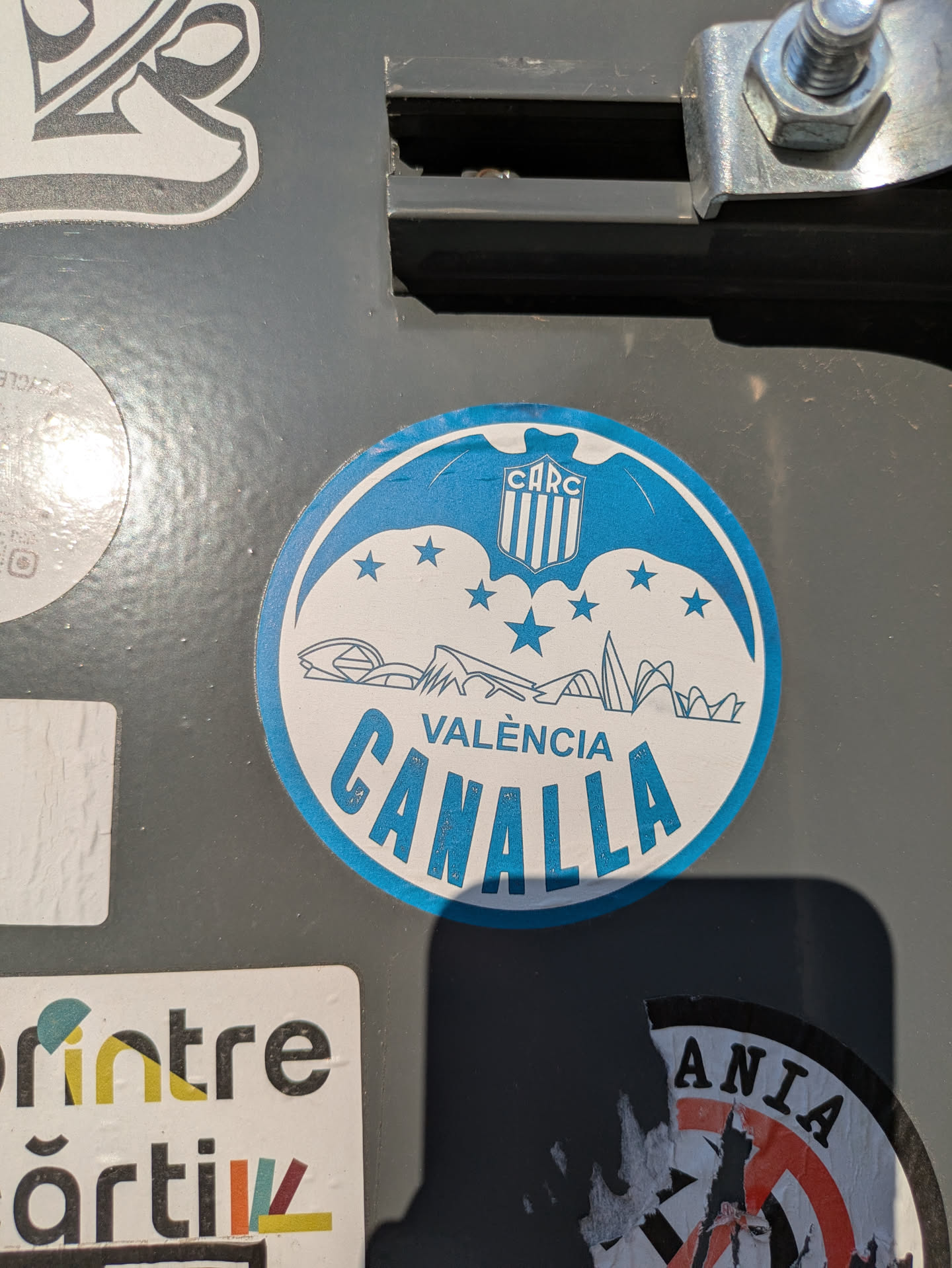 A round blue and white sticker features the text "VALENCIA CANALLA" in blue letters. Above the text is a stylized depiction of buildings and a logo with the letters "CARC". There are also several stars. The sticker appears to be adhered to a gray surface.