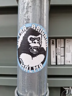 A sticker featuring a black and white illustration of a gorilla's head with the text "Ultra Brigada Malmö" around it. The sticker is placed on a metal pole.