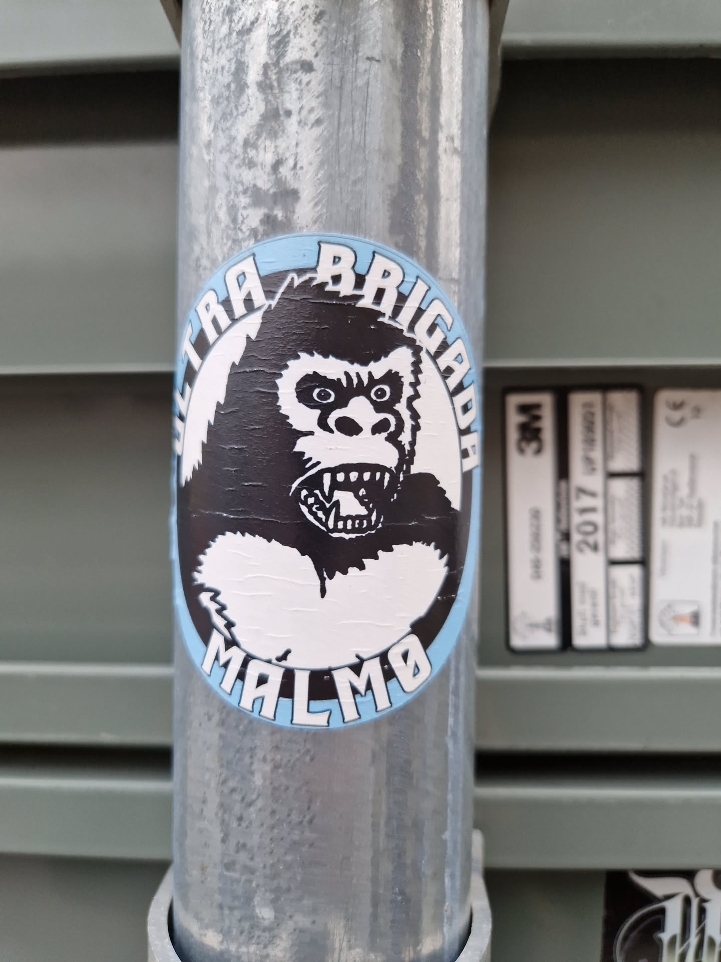 A sticker featuring a black and white illustration of a gorilla's head with the text "Ultra Brigada Malmö" around it. The sticker is placed on a metal pole.
