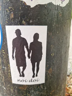 A sticker featuring a minimalist design of a man and a woman walking hand-in-hand. Their silhouettes are depicted against a white background, with the phrase "noi doi" written at the bottom.  The sticker is affixed to a dark-colored pole.