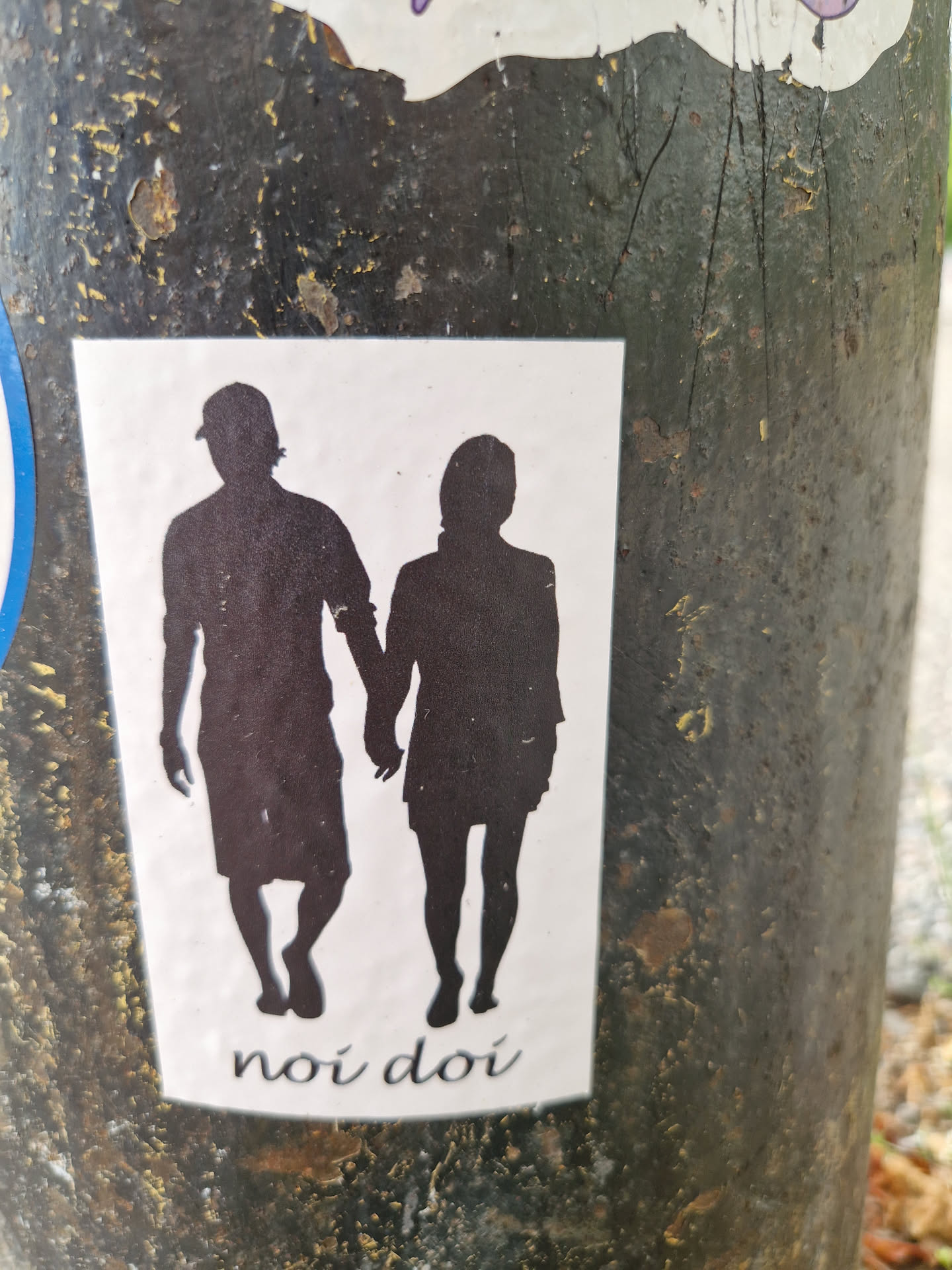 A sticker featuring a minimalist design of a man and a woman walking hand-in-hand. Their silhouettes are depicted against a white background, with the phrase "noi doi" written at the bottom.  The sticker is affixed to a dark-colored pole.