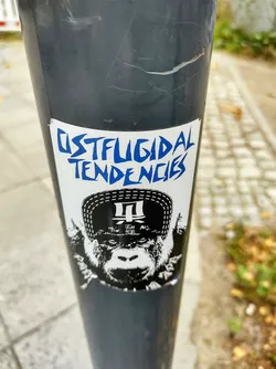 A sticker featuring a black and white illustration of a monkey wearing a hat. The text "OSTFLUGIDAL TENDENCIES" is written in blue above the monkey. 