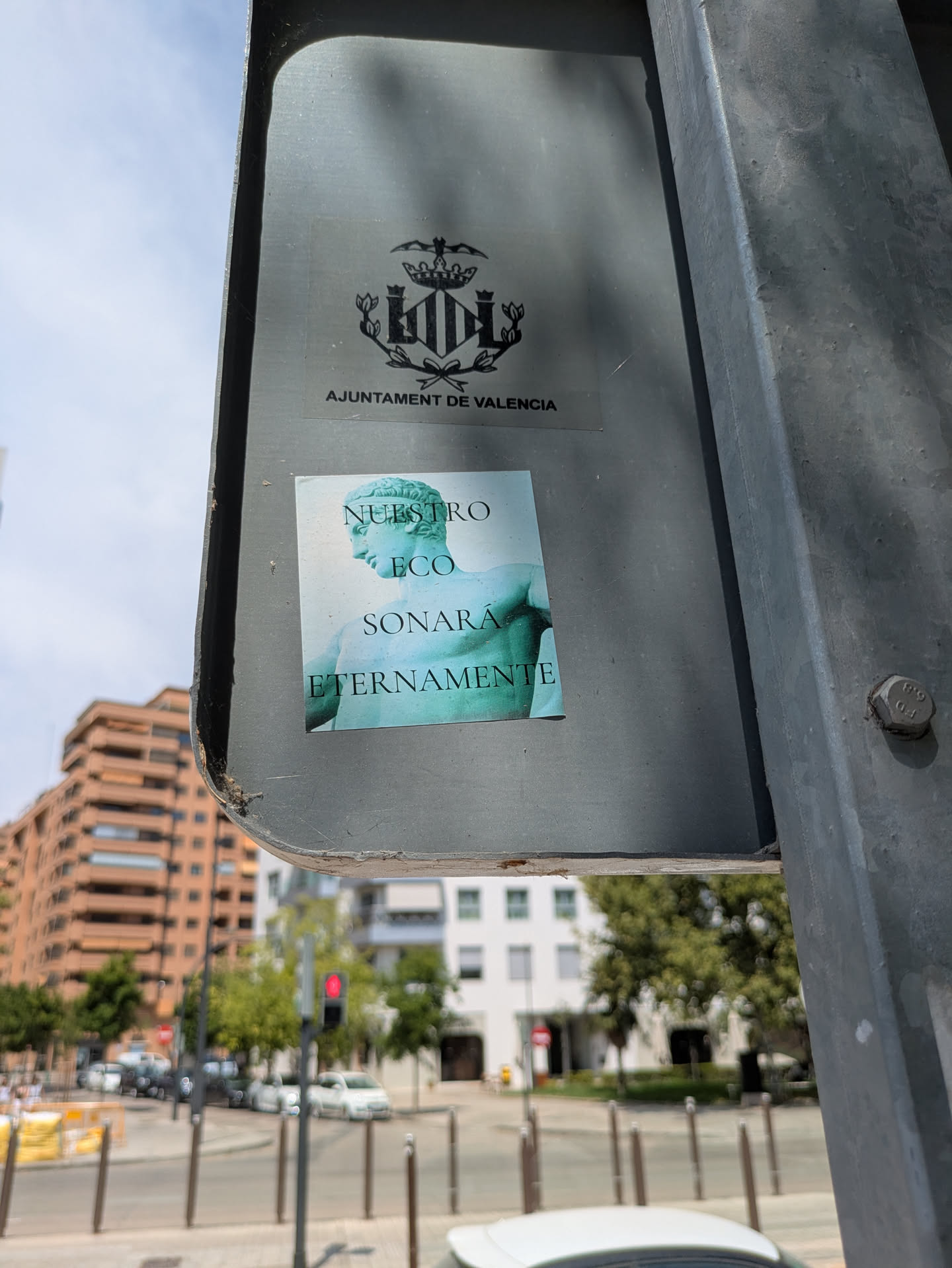 The sticker features a classical sculpture and the phrase "Nuestro eco sonará eternamente" which translates to "Our echo will sound eternally". The sign also has the coat of arms and the name of the city in catalan: Ajuntament de Valencia. The background shows an urban street scene during daytime.
