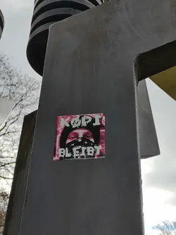 A sticker featuring a person wearing a mask, with the text "KOPI BLEIBT" (Kopi Remains) in stylized lettering. The overall color scheme is pink and white on a dark background. The sticker is affixed to a dark gray metal structure.