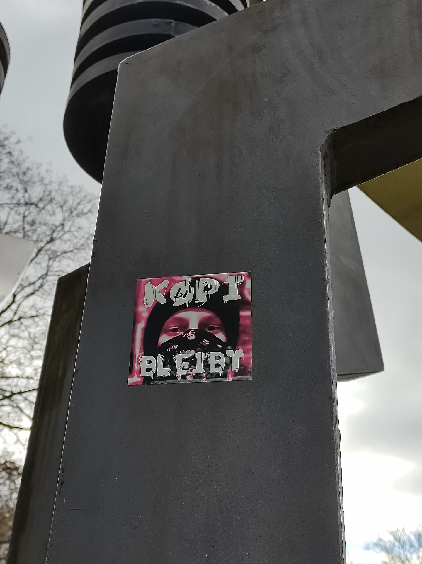 A sticker featuring a person wearing a mask, with the text "KOPI BLEIBT" (Kopi Remains) in stylized lettering. The overall color scheme is pink and white on a dark background. The sticker is affixed to a dark gray metal structure.