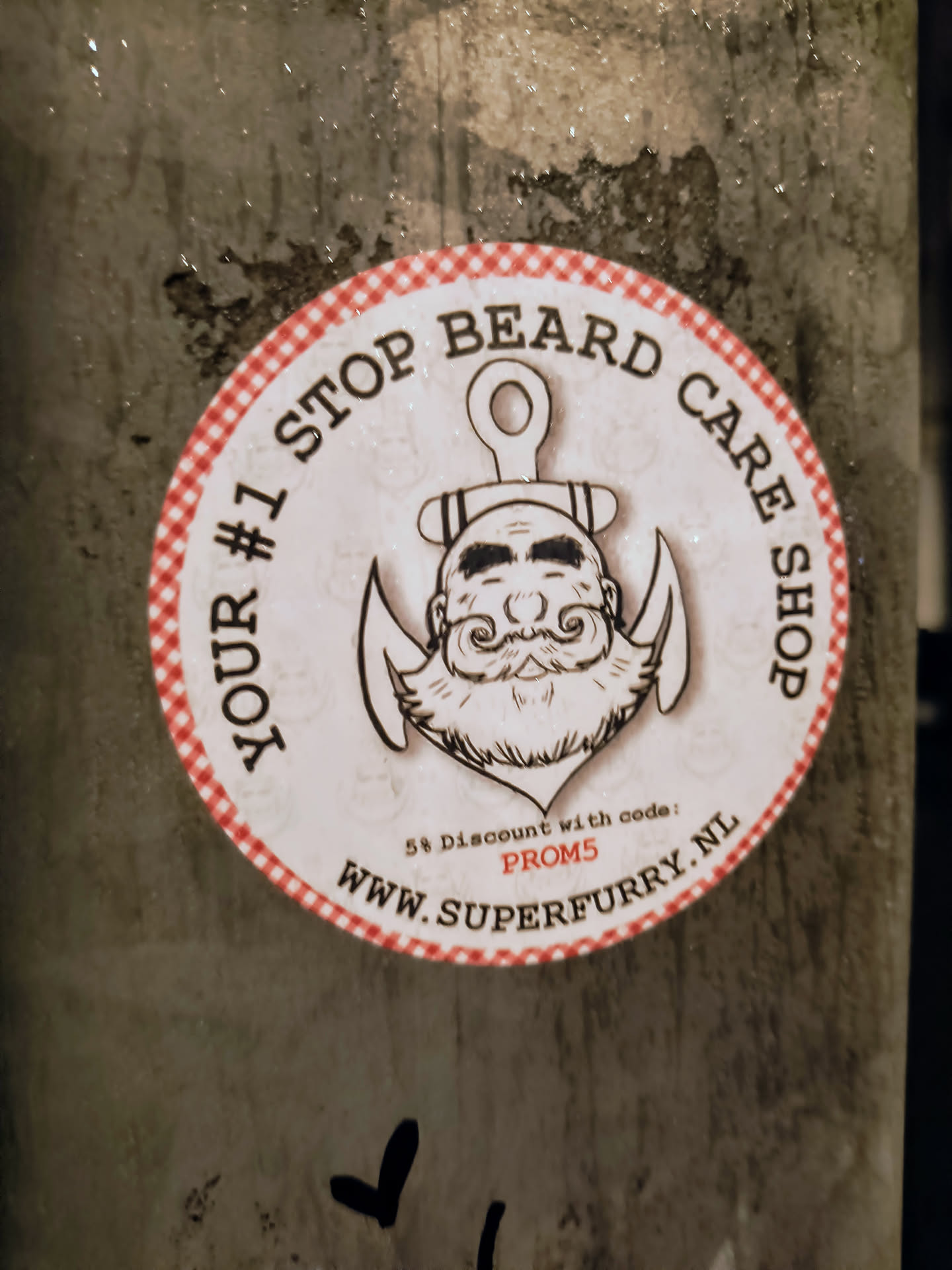 A round sticker with a red and white checkered border features an illustration of a bearded man with an anchor above his head, advertising "YOUR #1 STOP BEARD CARE SHOP". A promotional offer is mentioned at the bottom: "5% Discount with code: PROM5", along with the website address "WWW.SUPERFURRY.NL".