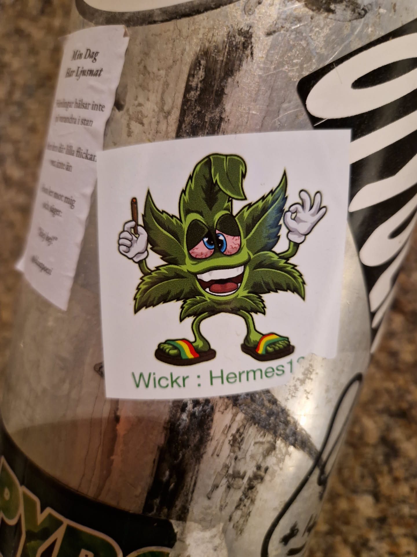 A sticker featuring a cartoon marijuana leaf character with red eyes, holding a joint in one hand and making the OK sign with the other. It's wearing rainbow sandals and has a happy expression. The text 'Wickr: Hermes10' is visible at the bottom.