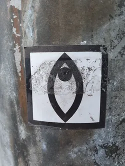 A black and white sticker featuring an abstract symbol resembling an eye. The symbol is enclosed in a square border. The sticker is affixed to a weathered gray surface.