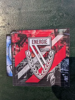 This is a square sticker featuring the Energie Cottbus logo, which is a stylized 'V' surrounded by a laurel wreath. The background consists of red and white stripes, images of fans, and what appears to be red smoke flares. The sticker looks like it's sitting on a textured surface.
