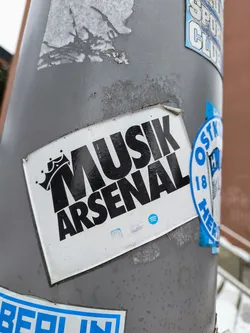 Black and white sticker with the text Musik Arsenal and a crown graphic.  There are also small social media icons visible at the bottom.