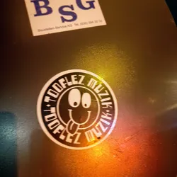 A round sticker with a smiley face and the words "Toooflez Muzik" written around it. The smiley face has big eyes and a tongue sticking out.