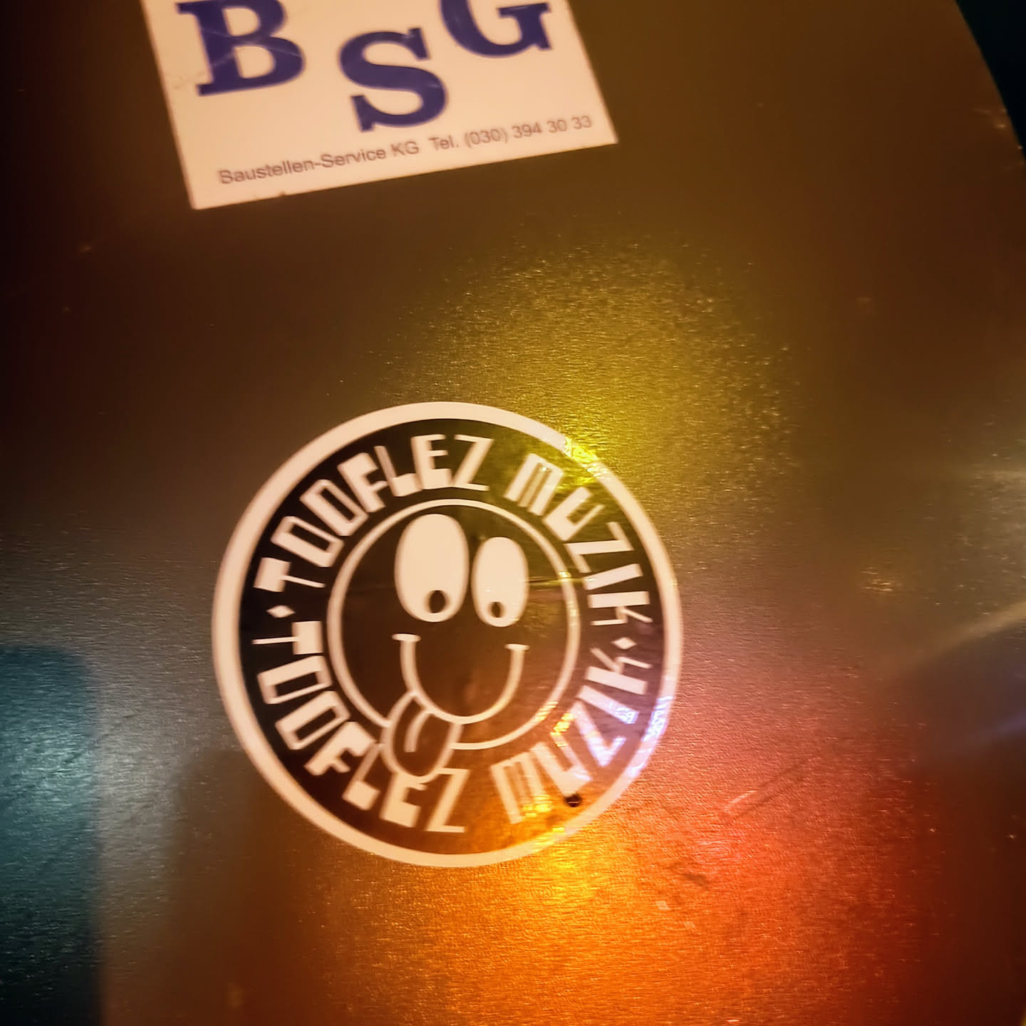 A round sticker with a smiley face and the words "Toooflez Muzik" written around it. The smiley face has big eyes and a tongue sticking out.