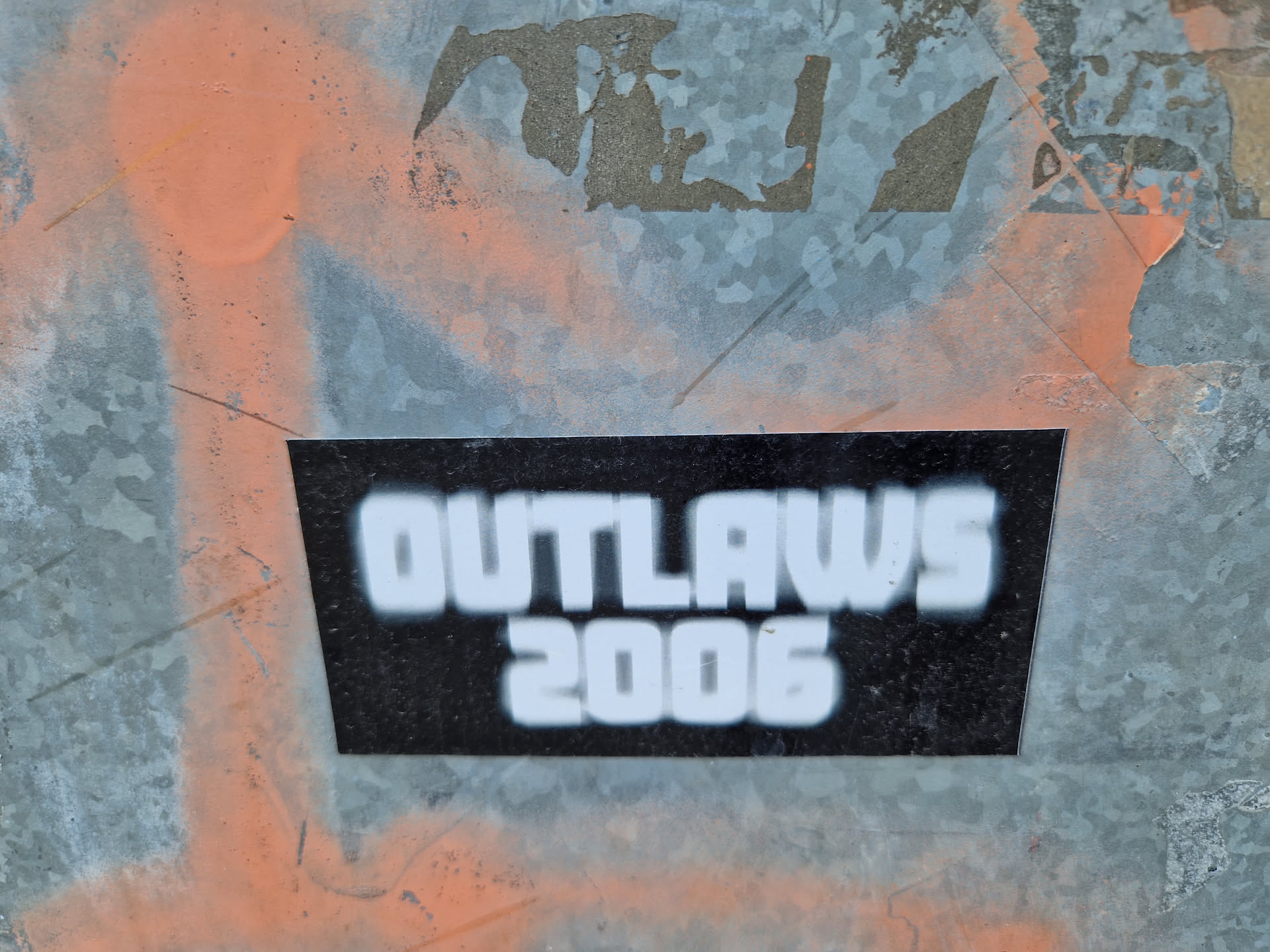 A black and white sticker with the text "OUTLAWS 2006" on it. The sticker is affixed to a metallic surface with some paint splatters on it.