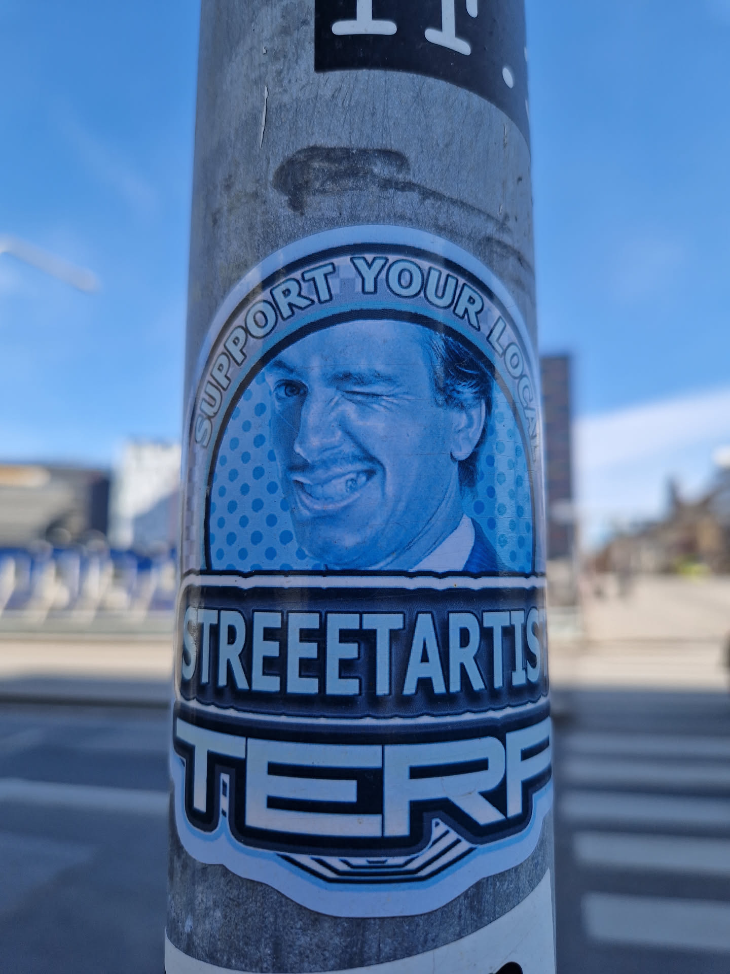 A blue and white sticker featuring a winking man's face in a circle with the text 'Support Your Local' above and 'STREETARTISTERF' below. The sticker has a pop art style.