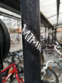 The sticker is rectangular, featuring the word "MIMMI" in a prominent, white, brushstroke-style font against a solid black background. The letters have a dynamic, hand-painted appearance with visible texture, suggesting a raw, energetic aesthetic. The sticker is applied diagonally on a dark, textured metal pole, contrasting sharply with its surroundings.