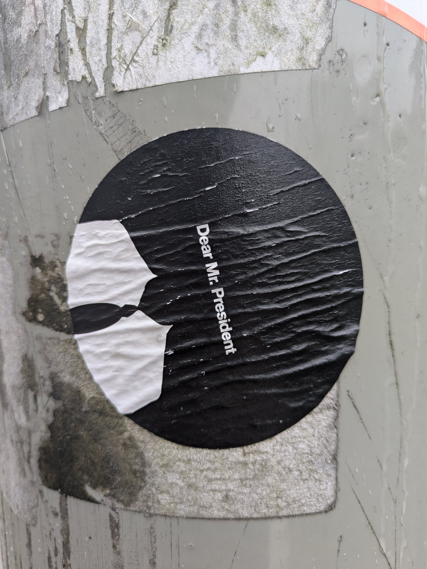 This round sticker features a stark contrast of black and white. The text 'Dear Mr. President' is printed along the right side of the sticker. To the left of the text is an abstract image of a broken white tie set against the black background. The sticker is adhered to a gray textured surface.