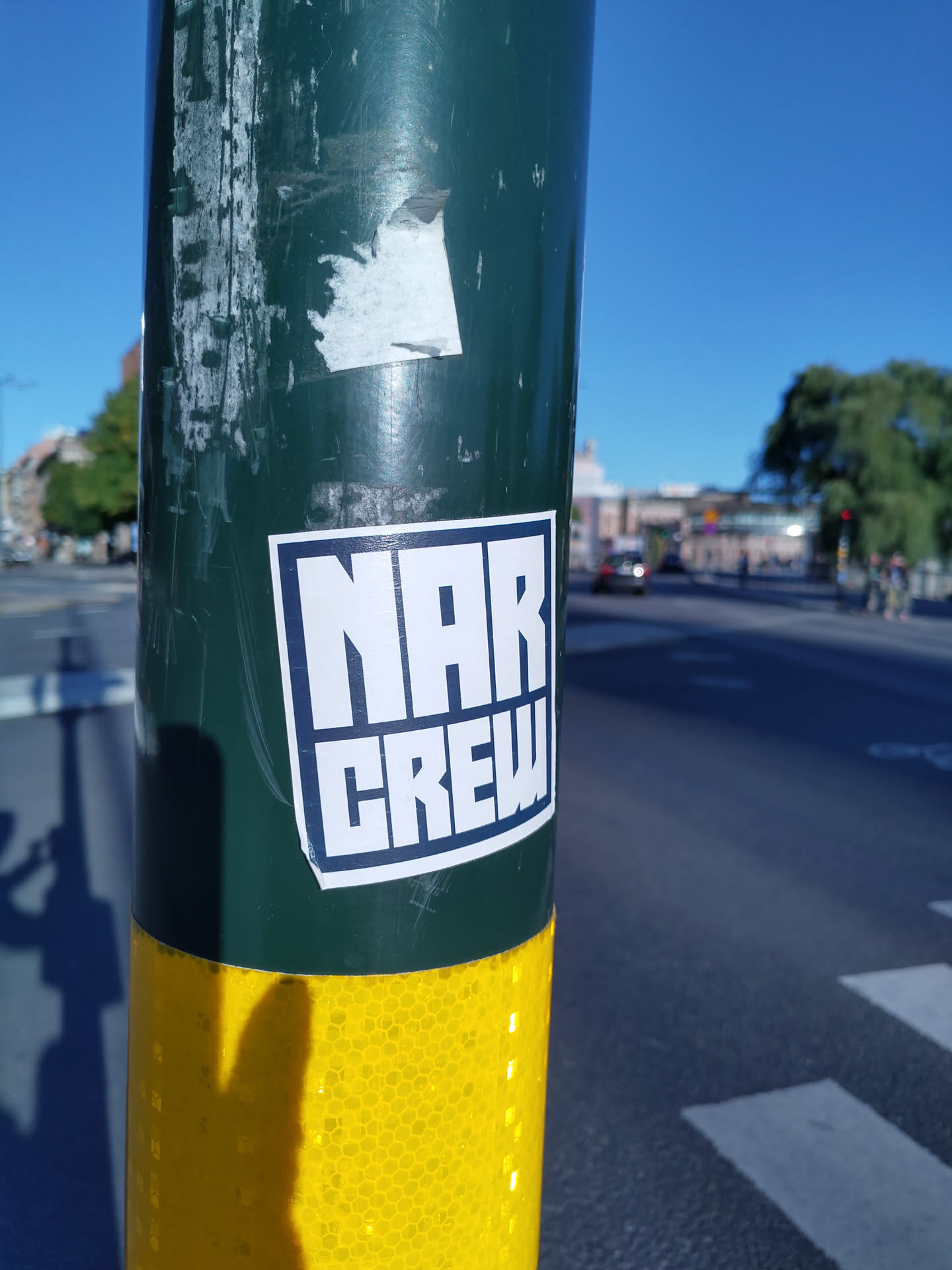 A close-up shot of a sticker on a pole. The sticker is predominantly white with black text that reads 'NAR CREW' in bold, uppercase letters.  The sticker is affixed to a dark green pole with a yellow reflective section at the bottom. The background shows a sunny city street scene.