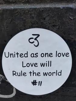 A round white sticker with a handwritten message promoting unity and love. It includes a small symbol resembling a stylized 'C3' at the top.