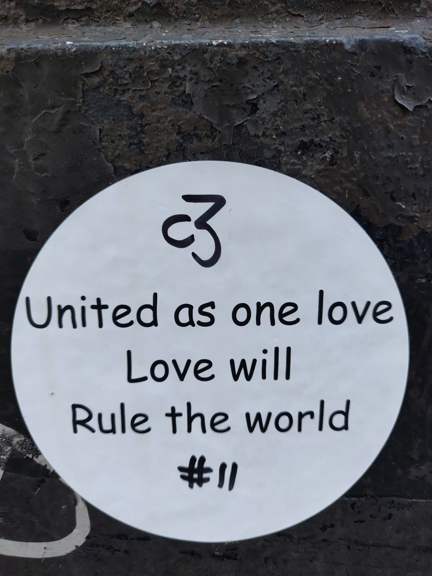 A round white sticker with a handwritten message promoting unity and love. It includes a small symbol resembling a stylized 'C3' at the top.