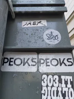A white sticker with the text PEOKSPEOKS in bold, uppercase, black letters. The sticker is slightly wet, and there are other stickers around it, including one that says "JÄER" and another circular sticker with illegible text and a logo.