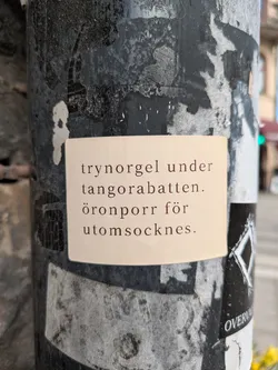 A beige rectangular sticker is adhered to a weathered pole, displaying text in Swedish. The pole has a dark, mottled appearance from accumulated grime and possible peeling paint. The text on the sticker reads "trynorgel under tangorabatten. öronporr för utomsocknes."