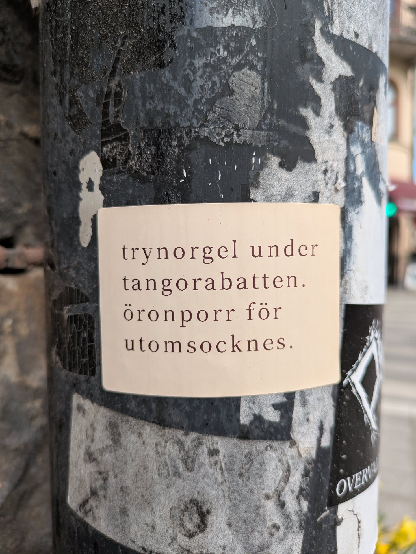 A beige rectangular sticker is adhered to a weathered pole, displaying text in Swedish. The pole has a dark, mottled appearance from accumulated grime and possible peeling paint. The text on the sticker reads "trynorgel under tangorabatten. öronporr för utomsocknes."