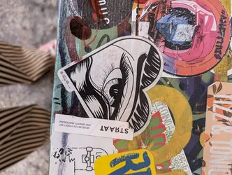 A black and white illustration of an eye forming a heart shape. The sticker has the artist's signature '@ealva_' and 'STRAAT' written at the bottom, suggesting it might be related to street art or urban art.  The sticker is affixed to a surface already covered with various other stickers and graffiti.