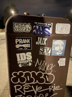 Street sticker A rectangular black sticker featuring the stylized graffiti name 'JAIK' in bold white block letters filled with vertical black stripes. In the lower right corner, the text 'V4E' is printed in smaller white block font. The sticker is placed on a brown utility box surrounded by other stickers and graffiti markings.