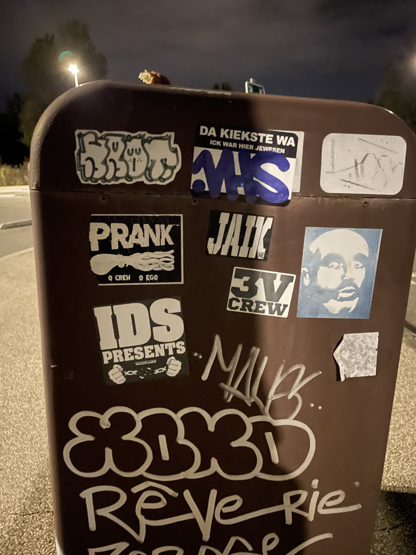A rectangular black sticker featuring the stylized graffiti name 'JAIK' in bold white block letters filled with vertical black stripes. In the lower right corner, the text 'V4E' is printed in smaller white block font. The sticker is placed on a brown utility box surrounded by other stickers and graffiti markings.