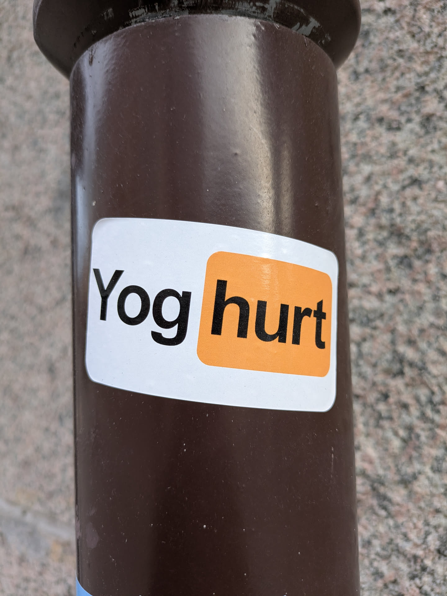 A close-up shot of a rectangular sticker placed on a dark brown pole. The sticker has a white background with the text "Yog hurt" written on it. The word "Yog" is printed in black, while the word "hurt" is inside an orange rounded rectangle, reminiscent of a popular website logo. This seems to be a play on words with the name of the website.
