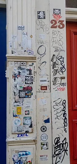 This image captures a corner of a building that's heavily decorated with various stickers and graffiti. The stickers range from simple designs to more elaborate artistic expressions, while the graffiti includes handwritten tags and abstract markings, creating a layered and textured appearance. The overall aesthetic suggests a vibrant urban environment where public spaces are used for self-expression and communication.