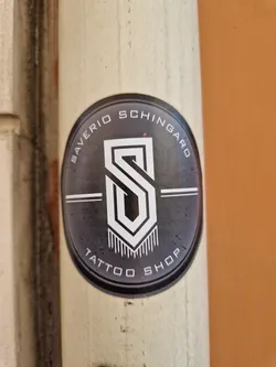 Oval sticker with a dark gray background and white text and logo. The logo is a stylized "S" inside a geometric shape, with vertical lines at the bottom. The text around the logo says "SAVERIO SCHINGARO TATTOO SHOP".