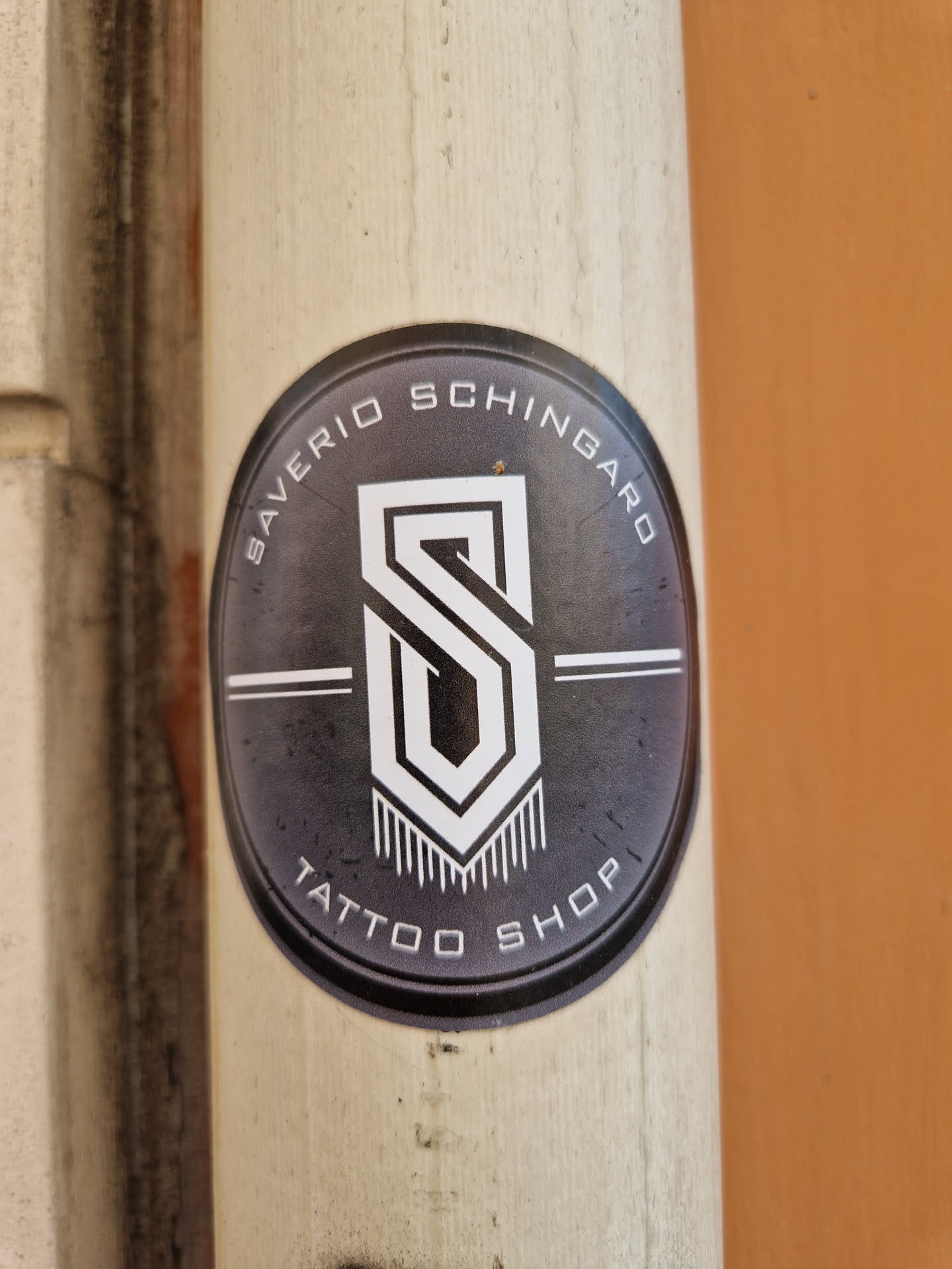 Oval sticker with a dark gray background and white text and logo. The logo is a stylized "S" inside a geometric shape, with vertical lines at the bottom. The text around the logo says "SAVERIO SCHINGARO TATTOO SHOP".