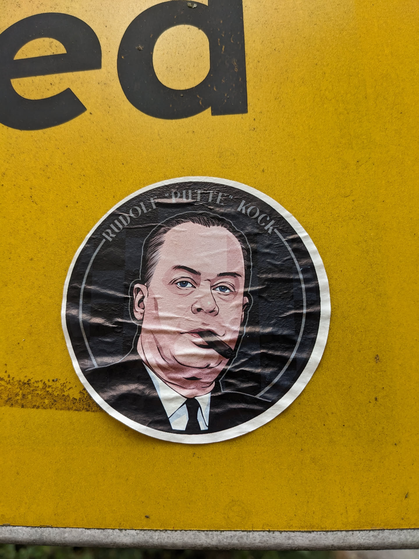 The sticker is circular and features a black and white illustration of a man's head and shoulders. He has a prominent mustache, short dark hair, and is wearing a suit and tie. The illustration is detailed with shading to give it depth. The man's face is rendered in pink and peach tones, with blue eyes. The background within the circle is black, and a thin white border surrounds the illustration. The sticker has visible creases and wrinkles, suggesting it has been applied to a surface for some time.