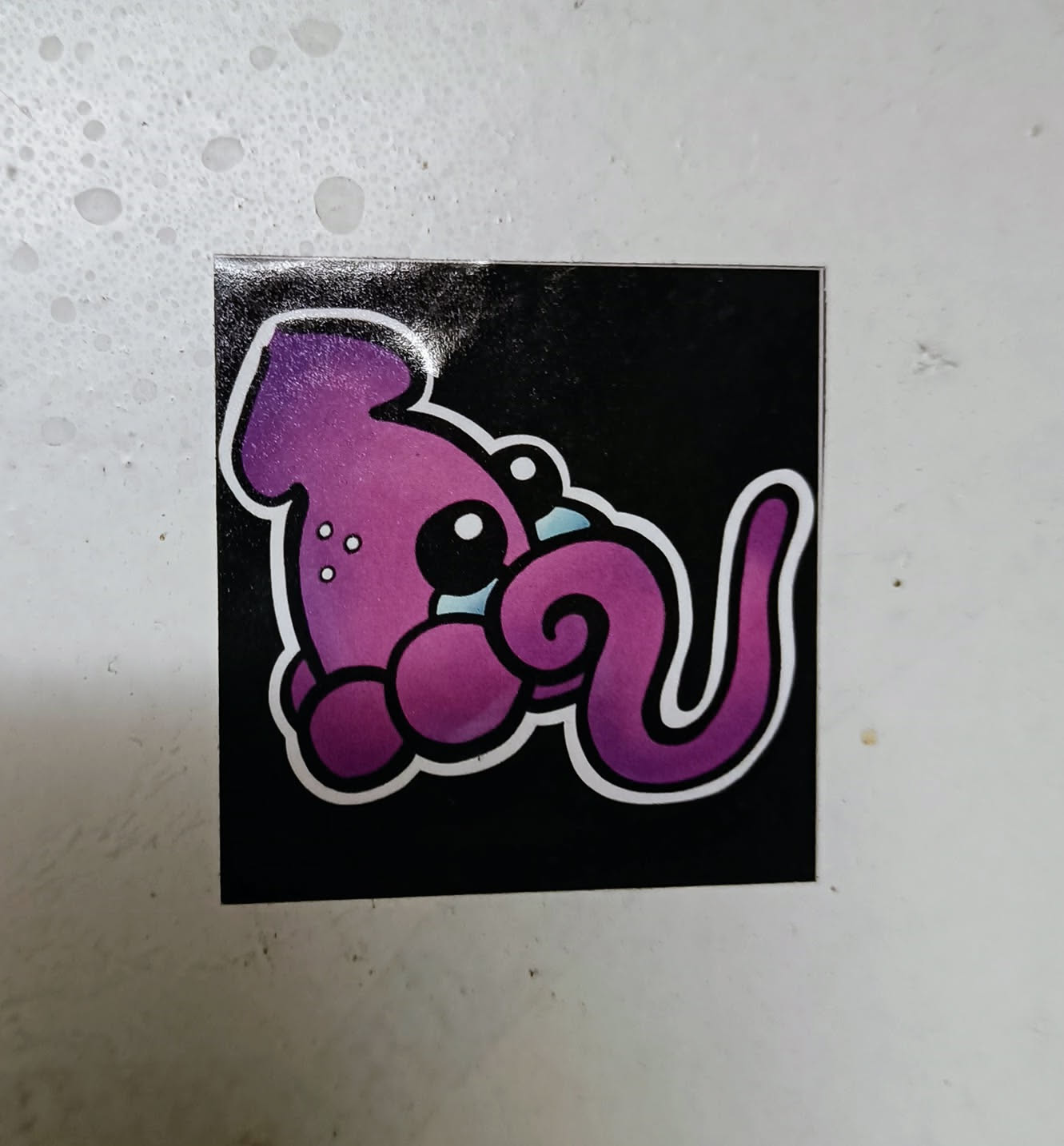 A cute purple squid with big eyes and a curled tentacle. The sticker has a black background and is printed with high quality.