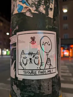 A white sticker is on a green pole, the sticker contains a drawing of a cat with a sad expression. Beside it, there is a bee, a balloon with a sad face and a ghost. The text on the sticker says "TROUBLE FINDS ME!".