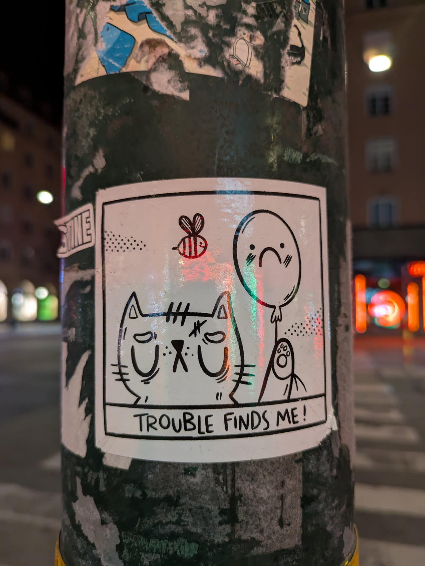 A white sticker is on a green pole, the sticker contains a drawing of a cat with a sad expression. Beside it, there is a bee, a balloon with a sad face and a ghost. The text on the sticker says "TROUBLE FINDS ME!".