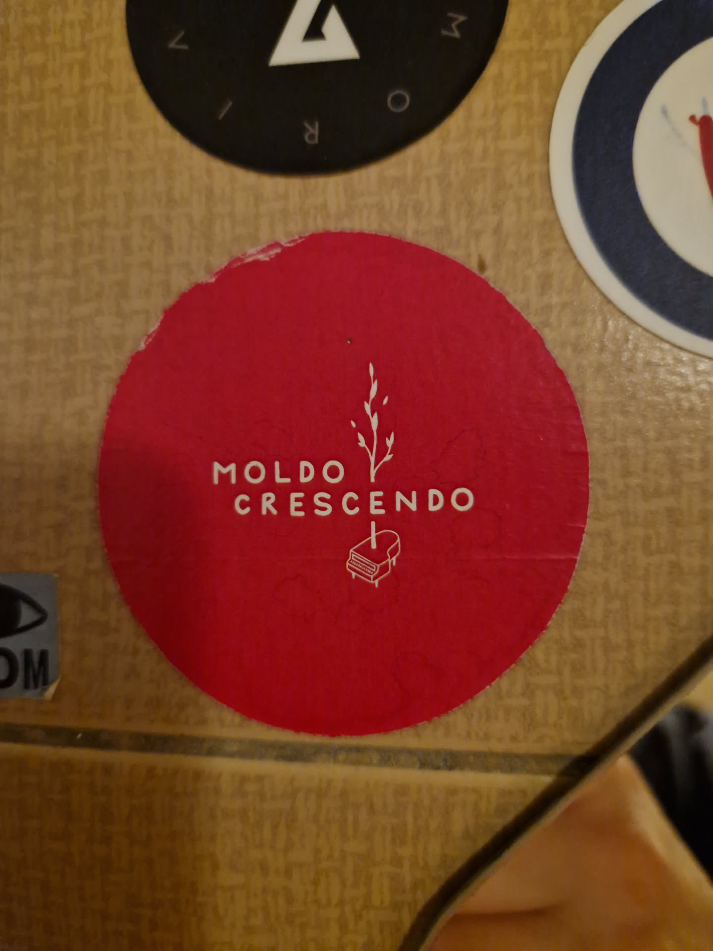 Red round sticker with the text "MOLDO CRESCENDO" in white, and a minimalist drawing of a plant and a piano.