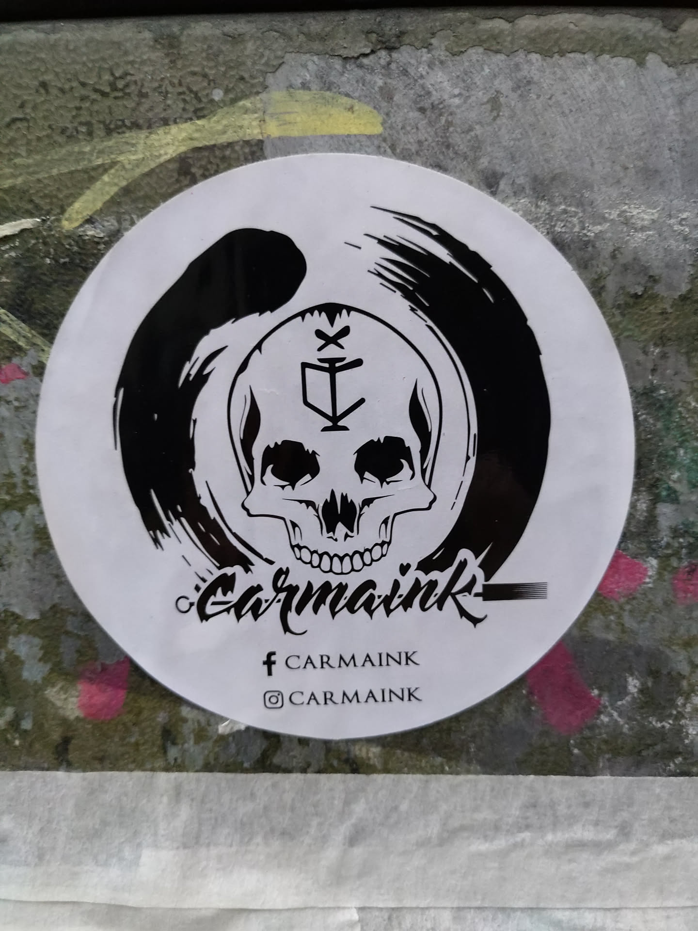 Round white sticker with a black and white illustration of a skull inside an enso circle. The skull has a symbol on its forehead.  Below the artwork, the text 'Carmaink' is written in a stylized script font. Social media handles for Facebook and Instagram are also present.