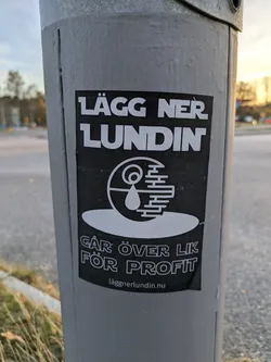 A black and white sticker with a design resembling the Death Star with a drop of blood in the center. The text on the sticker says "Lägg ner Lundin" (Shut down Lundin) and "Går över lik för profit" (Walks over corpses for profit) in Swedish. There is also a website address at the bottom: laggnerlundin.nu.