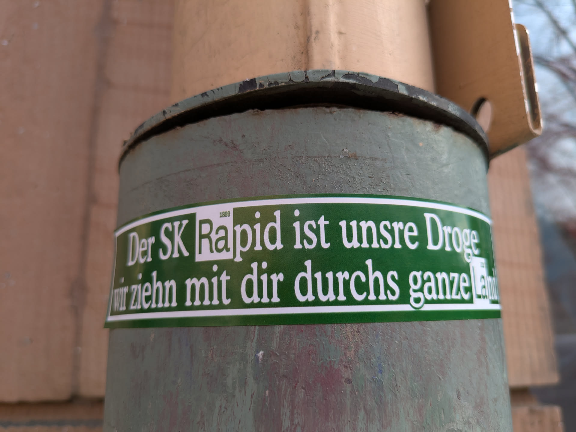 A green and white sticker wrapped around a pole, stating "Der SK Rapid ist unsere Droge, wir ziehn mit dir durchs ganze Land", in German. It translates to "SK Rapid is our drug, we'll move with you through the whole country".