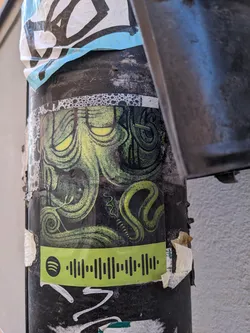 A sticker featuring a creepy, green creature with glowing eyes. It has a serpentine design element and includes a Spotify QR code at the bottom. The sticker is affixed to a dark-colored pole with other graffiti and stickers around it.