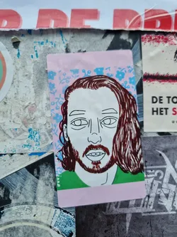 Close-up of a sticker on a wall. The sticker shows a line drawing of a man's face with long brown hair. The background is a light pink with a pattern of small blue and light green flowers. The man is wearing a green shirt. The style is simple, almost minimalist, with bold lines and flat colors.