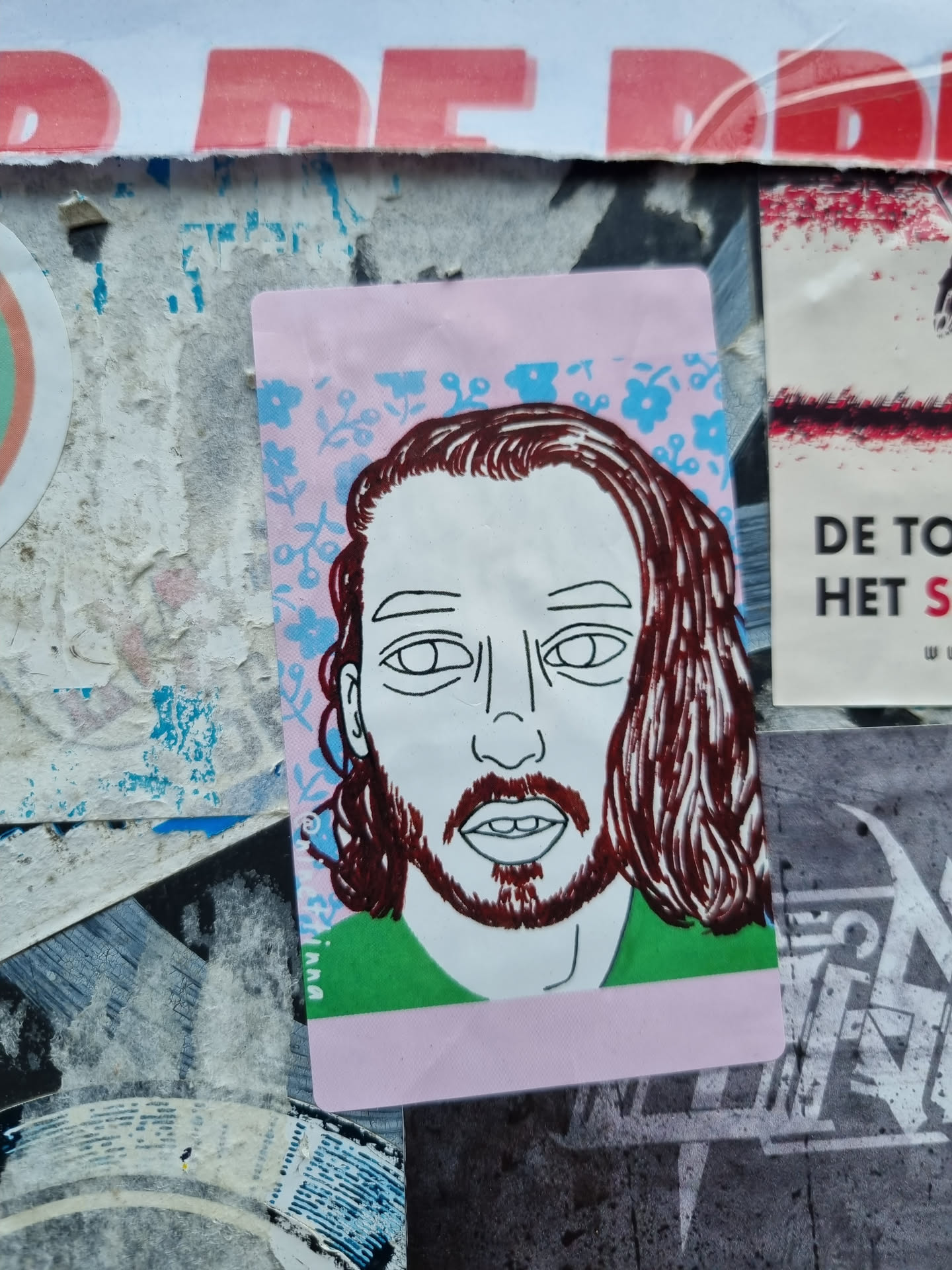 Close-up of a sticker on a wall. The sticker shows a line drawing of a man's face with long brown hair. The background is a light pink with a pattern of small blue and light green flowers. The man is wearing a green shirt. The style is simple, almost minimalist, with bold lines and flat colors.