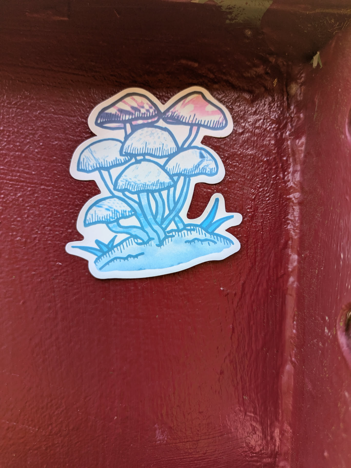 The sticker features an artistic illustration of several mushrooms growing together. The design uses watercolor-like effects with a light blue base for the stalks and lower caps. The top mushrooms show faint washes of pink and purple on their caps, creating a soft, ethereal look. All elements are outlined with a dark blue line. The sticker is placed on a rough, reddish-brown surface, possibly a metal pole or wall.