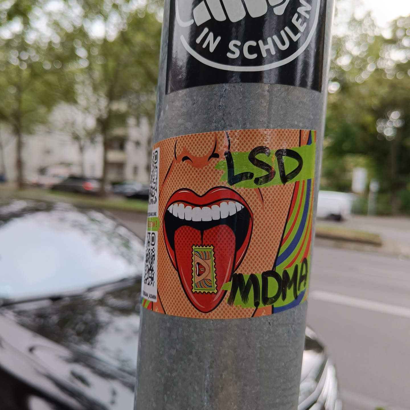 A sticker featuring a pop art style illustration of a woman's mouth with her tongue sticking out. The tongue has an LSD and MDMA stamp on it. The sticker has a QR code in the corner.