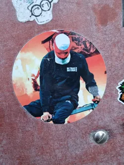 Round sticker featuring a stylized image of a person wearing a balaclava riding a bicycle in front of a fiery background. The person is wearing a black sweatshirt with the text "ULTRAS AMSTERDAM" visible on the chest. The overall style is bold and graphic.