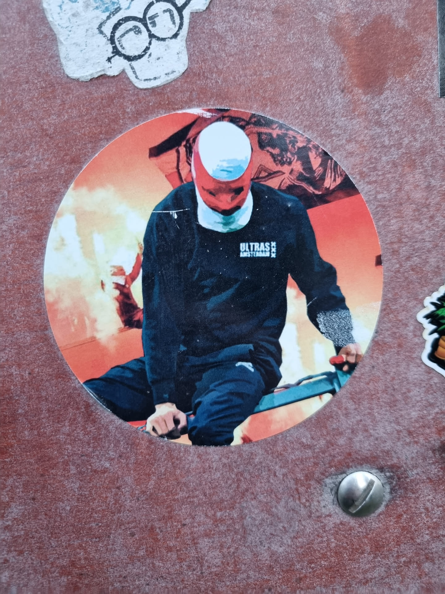 Round sticker featuring a stylized image of a person wearing a balaclava riding a bicycle in front of a fiery background. The person is wearing a black sweatshirt with the text "ULTRAS AMSTERDAM" visible on the chest. The overall style is bold and graphic.