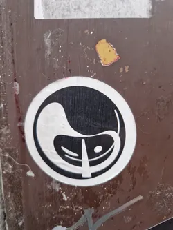 A circular sticker featuring a design reminiscent of the Yin and Yang symbol, but with a unique twist. The shapes are somewhat bean-like and incorporate a central line. It's adhered to a dark brown surface, possibly metal or wood, showing some wear and tear.
