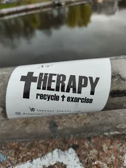 A sticker with a cross and the word THERAPY. Below it says 'recycle + exorcise'. It includes a website and Instagram account.